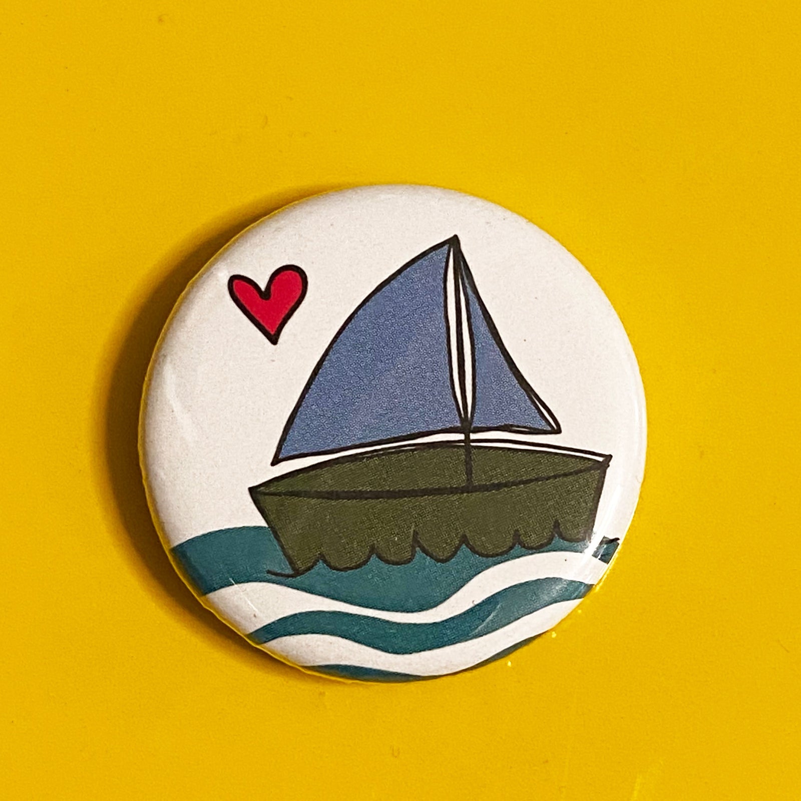 A round magnet with an illustration of a sailboat and a red heart, against a yellow backdrop 
