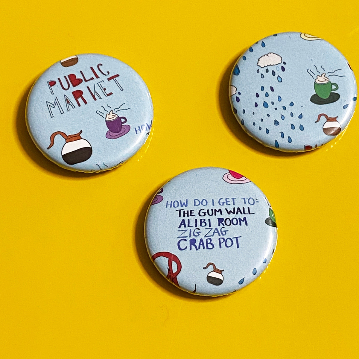 Three light blue magnets featuring coffee  cup and raining cloud drawings, and text that says "Public Market" and "How do I get to: the gum wall, Alibi Room, Zig Zag, Crab Pot" against a yellow background. 