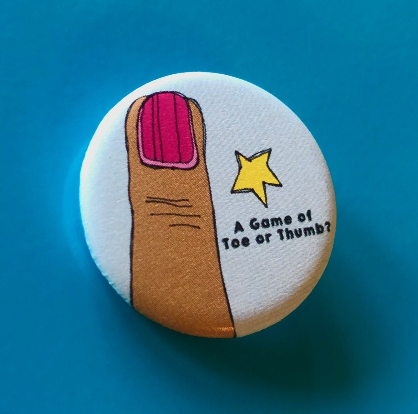 A round light blue magnet with a drawing of a finger with pink nail polish, a yellow star, and text that says "A Game of Toe or Thumb?" against a blue background. 