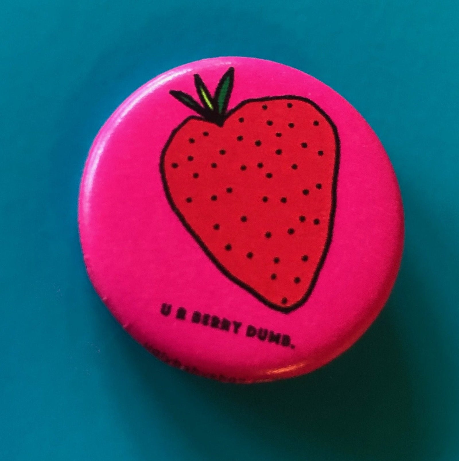 A round hot pink magnet with a drawing of a strawberry and text that says "U R Berry Dumb" against a blue background. 