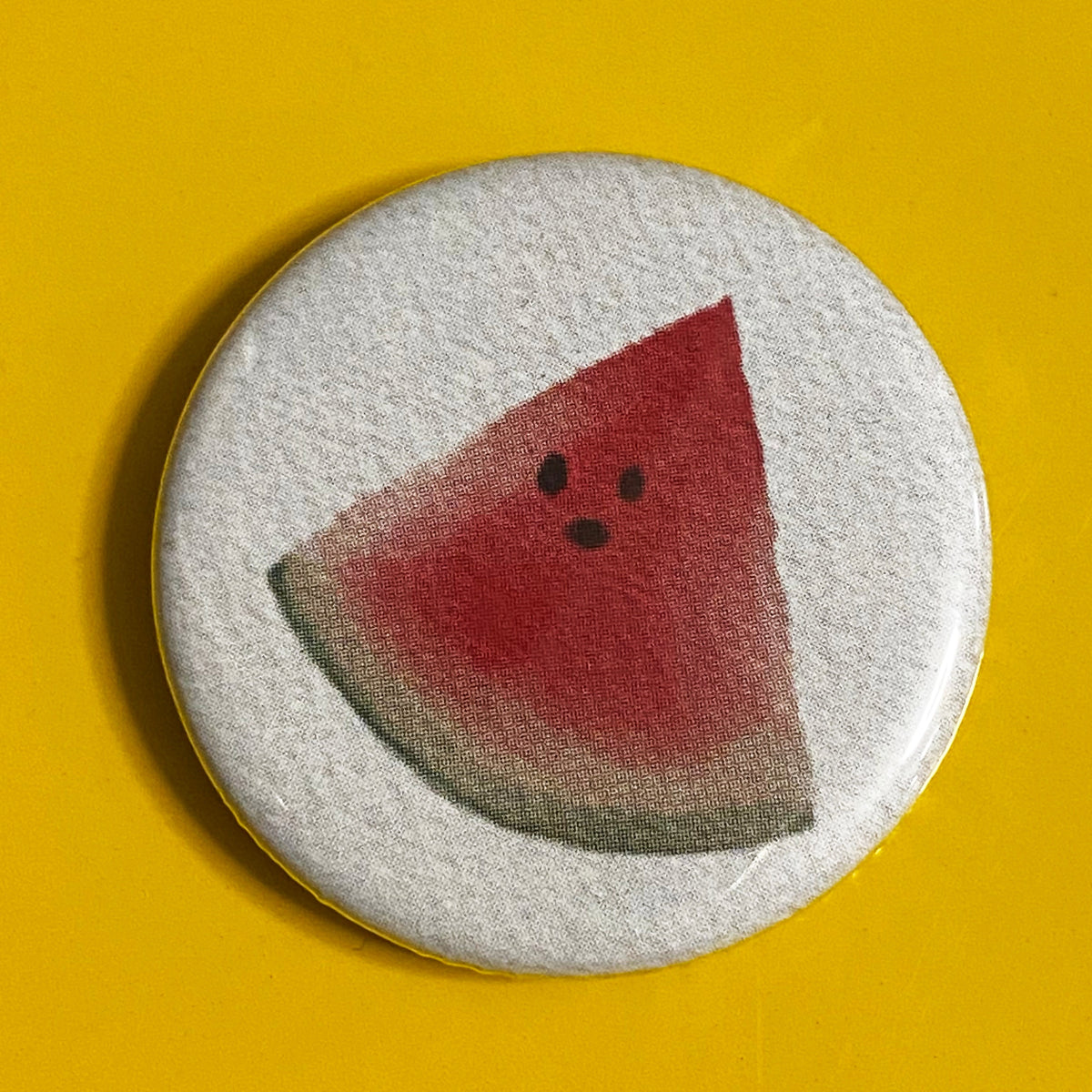 A round white magnet with an illustration of a slice of watermelon against a yellow background. 