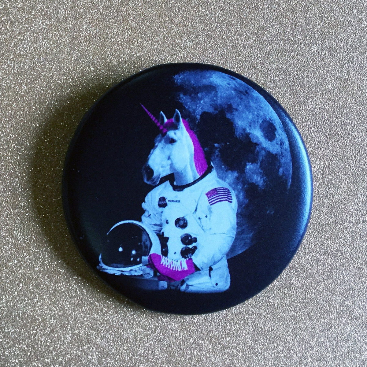 Round black magnet with an astronaut unicorn wearing roller skates with the moon in the background on top of a glittery gold backdrop. 