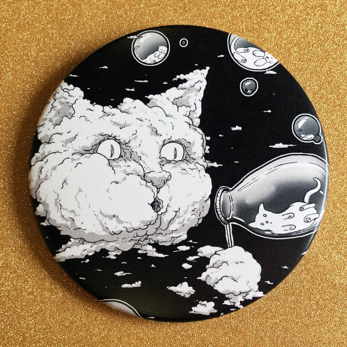 A round magnet with a black and white illustration of a cat-shaped cloud blowing bubbles that have little cats inside of them, on top of a gold glittery background. 