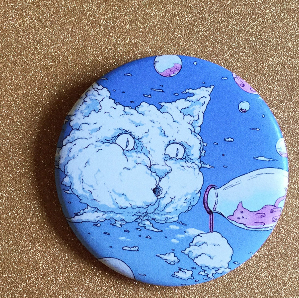 A blue magnet with an illustration of a cat-shaped cloud blowing bubbles that have little cats inside of them, against a glittery gold backdrop. 