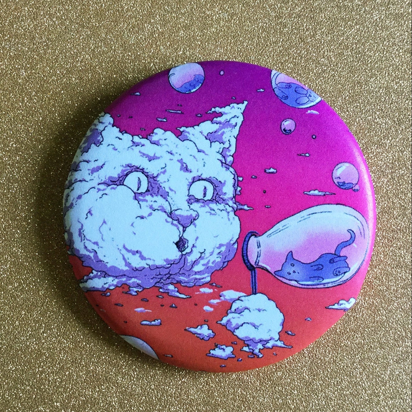 A round pink magnet with an illustration of a cat-shaped cloud blowing bubbles with little cats inside of them, against a glittery gold backdrop. 
