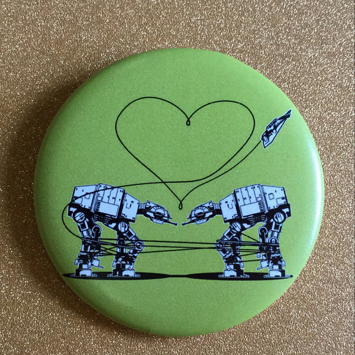 Round green magnet with an illustration of two AT-ATs from Star Wars with a heart above them, against a glittery gold background. 