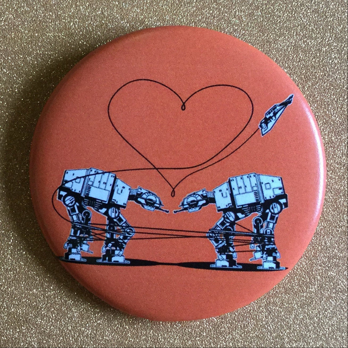 Round orange magnet with an illustration of two AT-ATs with a heart above them, against a gold glittery background. 