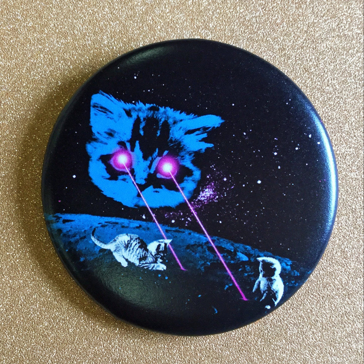 Round black magnet with an illustration of a large kitten shooting lasers out of its eyes that two smaller kittens are playing with. 