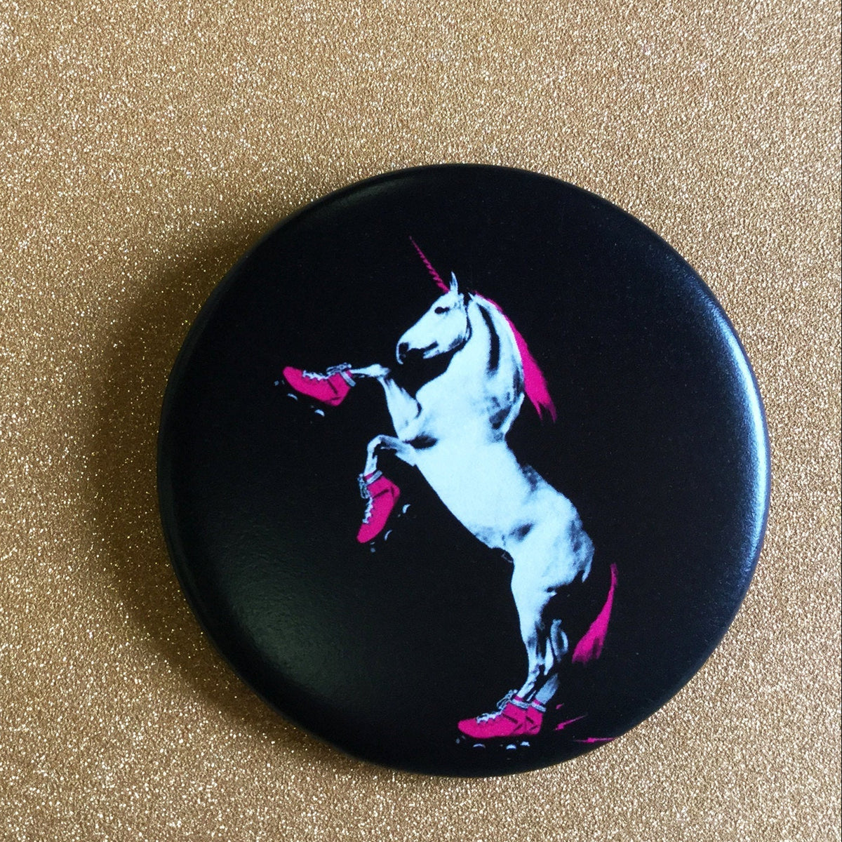 Round black magnet with an illustration of a unicorn with a pink mane and tail and rollerskates, against a glittery gold background. 