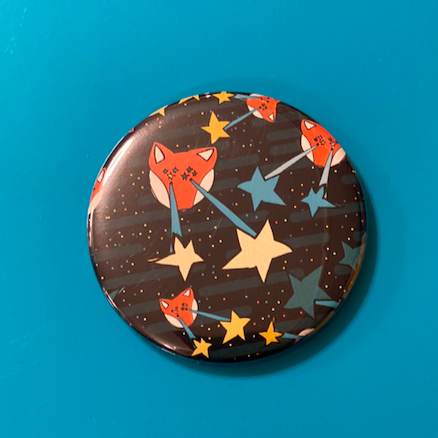 Round magnet with illustrations of foxes shooting stars out of their eyes, against a bright blue backdrop. 