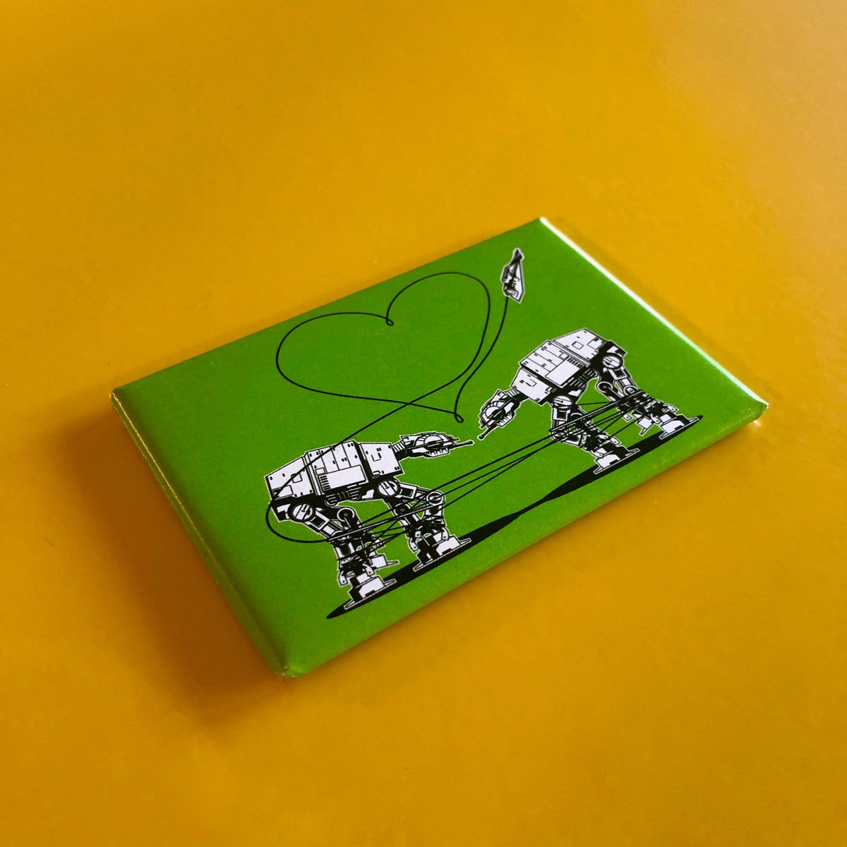 Green rectangular magnet with an illustration of two AT-ATs from Star Wars with a heart above them, against a yellow backdrop. 