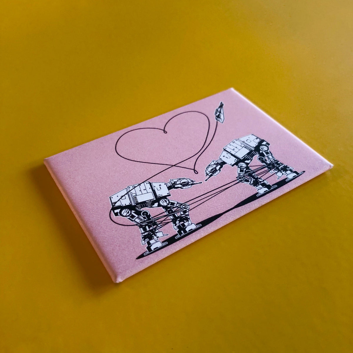 A pink rectangular magnet with an illustration of two AT-ATs from Star Wars with a heart above them, against a yellow backdrop. 