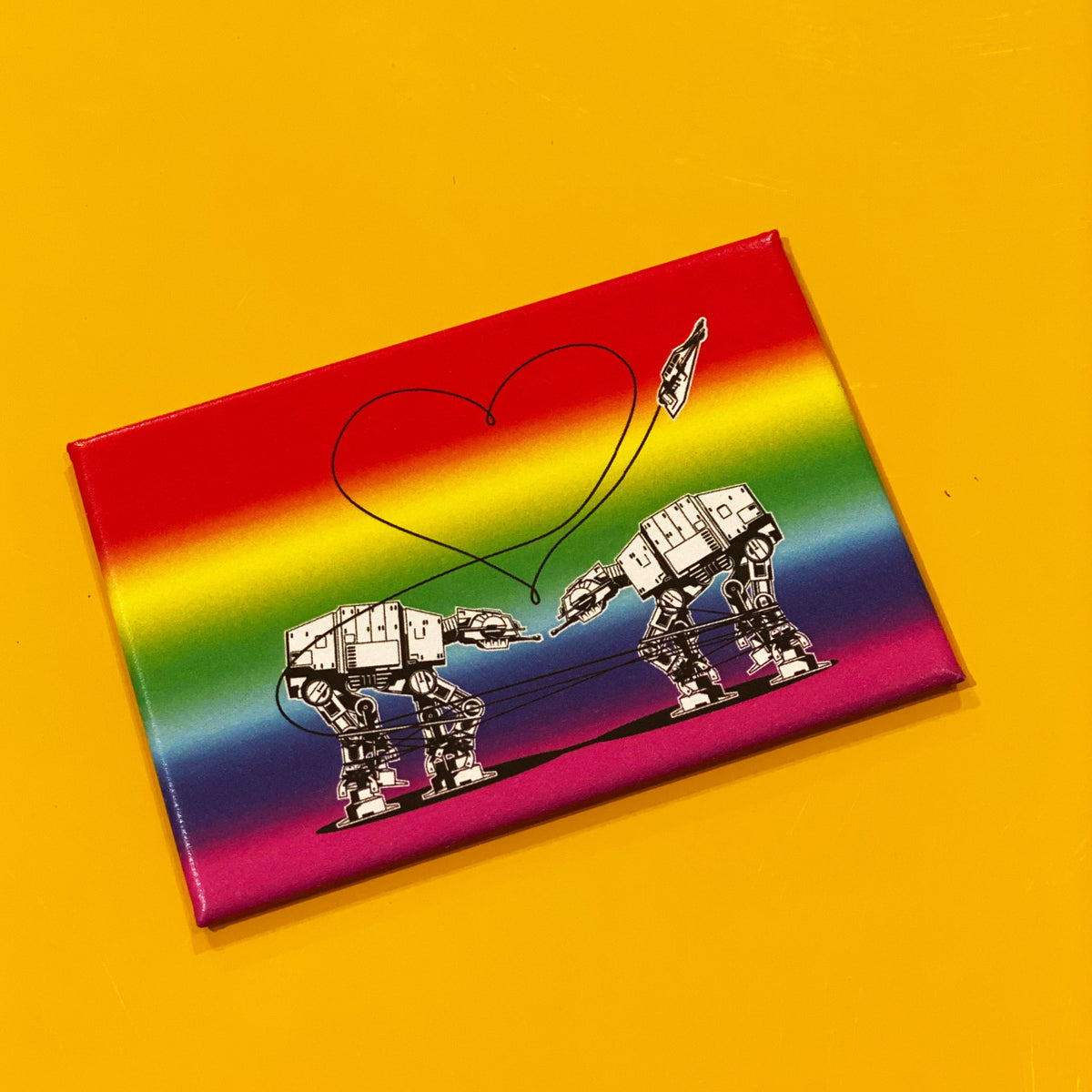 Rainbow rectangular magnet with an illustration of two AT-ATs from Star Wars with a heart above them, against a yellow backdrop. 