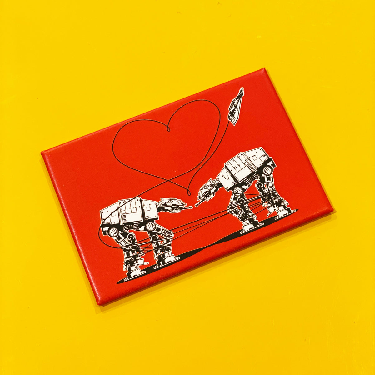 Red rectangular magnet with an illustration of two AT-ATs from Star Wars with a heart above them, against a yellow backdrop. 