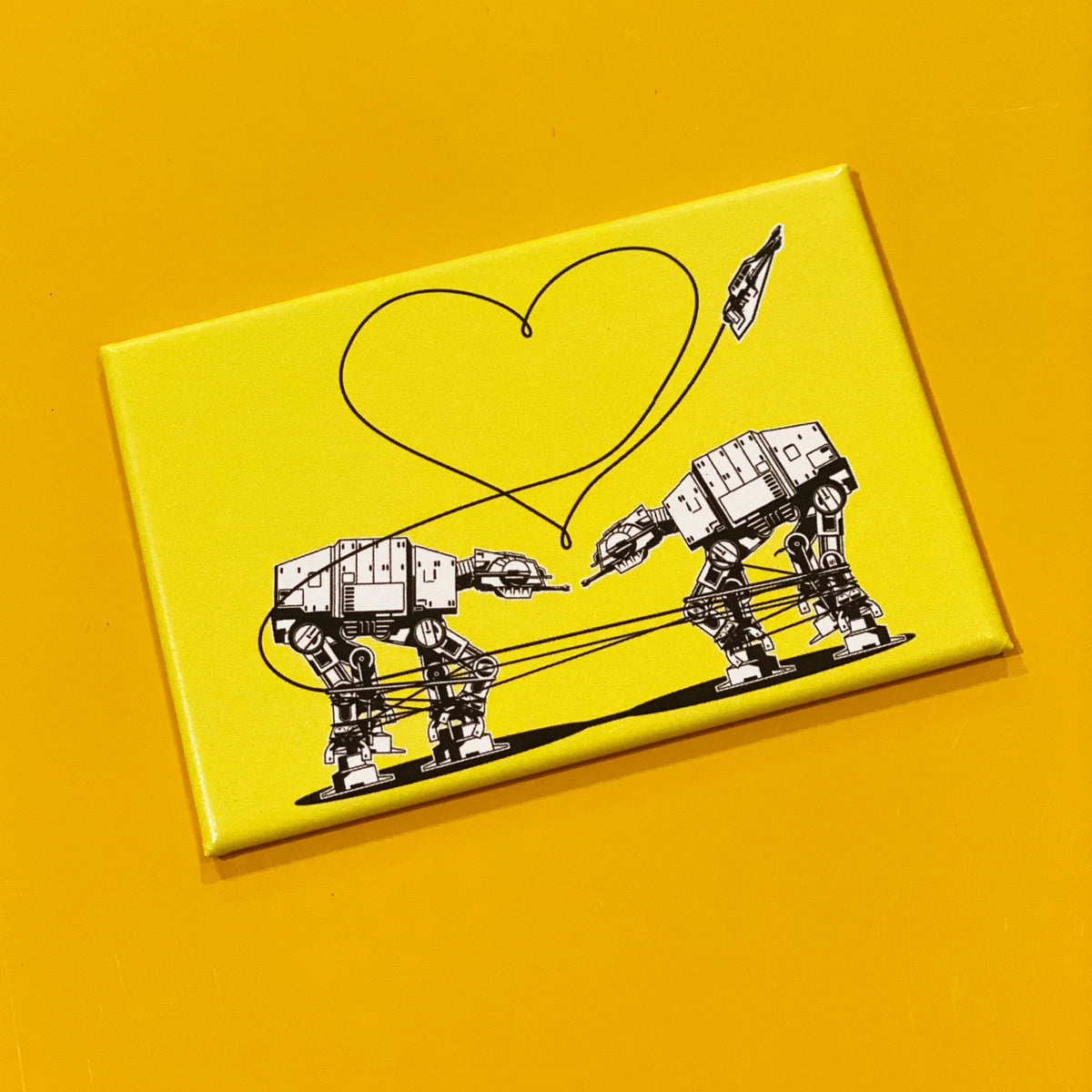 A yellow rectangular magnet with an illustration of two AT-ATs from Star Wars with a heart above them, against a darker yellow backdrop. 