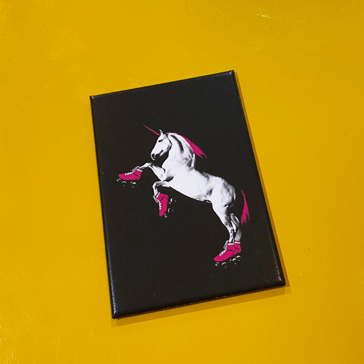 A black rectangular magnet with an illustration of a white unicorn with a pink mane and tail wearing pink roller skates. 
