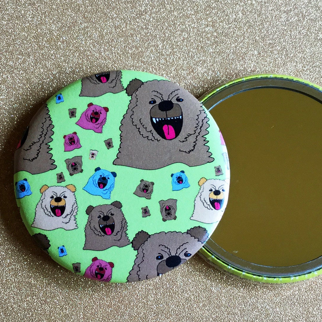 Two mirrors next to each other on a glittery gold surface. One is face up, showing its reflective surface, and the other is face down, showing its decorative back which has illustrations of cartoon bears on a green background. 
