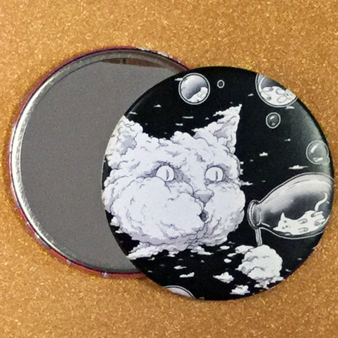 Two mirrors on a glittery gold surface, one face up showing its reflective side, and one face down showing its decorative back which has a black and white illustration of a cat-shaped cloud blowing bubbles with smaller cats inside of them. 