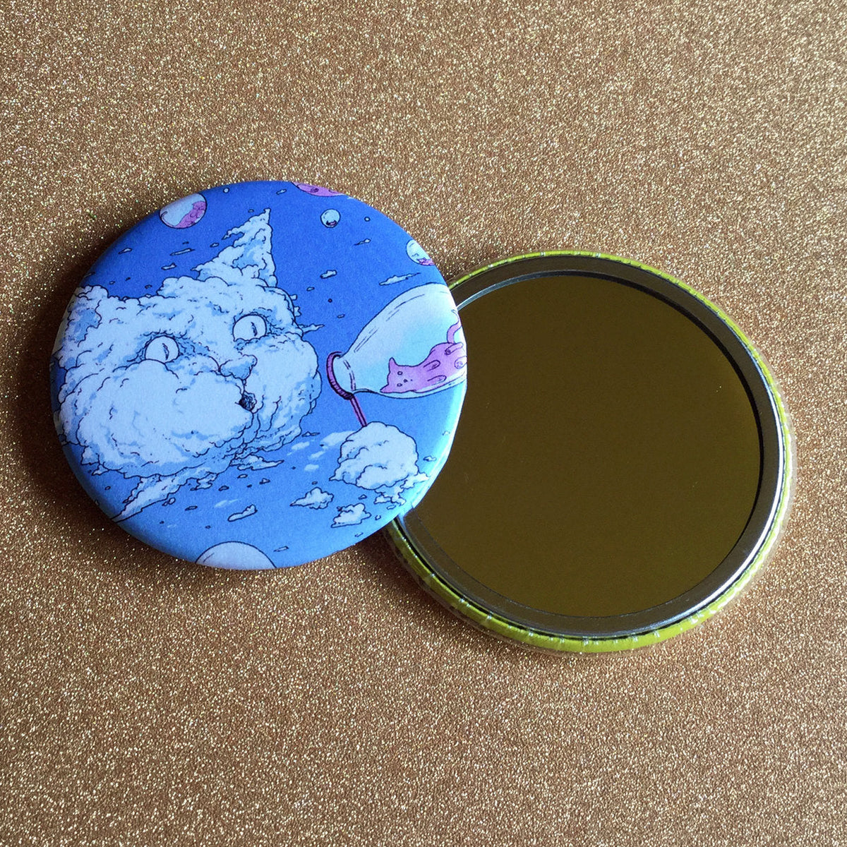 Two mirrors on a gold glittery background; one face up, showing its reflective surface, and the other face down, showing its decorative back, which has a blue illustration of a cat-shaped cloud blowing bubbles that have smaller cats inside of them. 