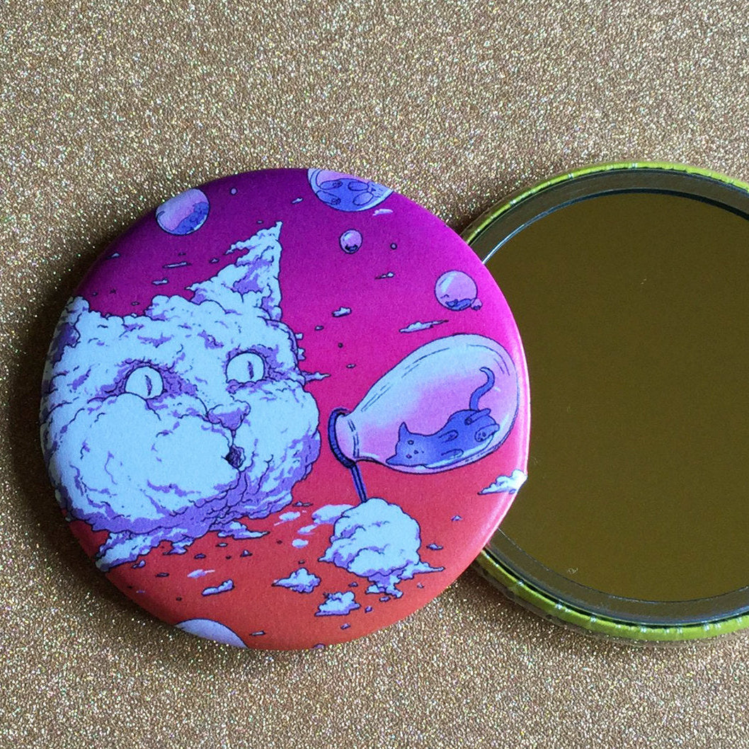 Two mirrors next to each other on a gold glittery surface, one face up showing its reflective surface, the other face down showing its decorative back, which has a pink illustration of a cat-shaped cloud blowing bubbles that have tiny cats inside of them. 