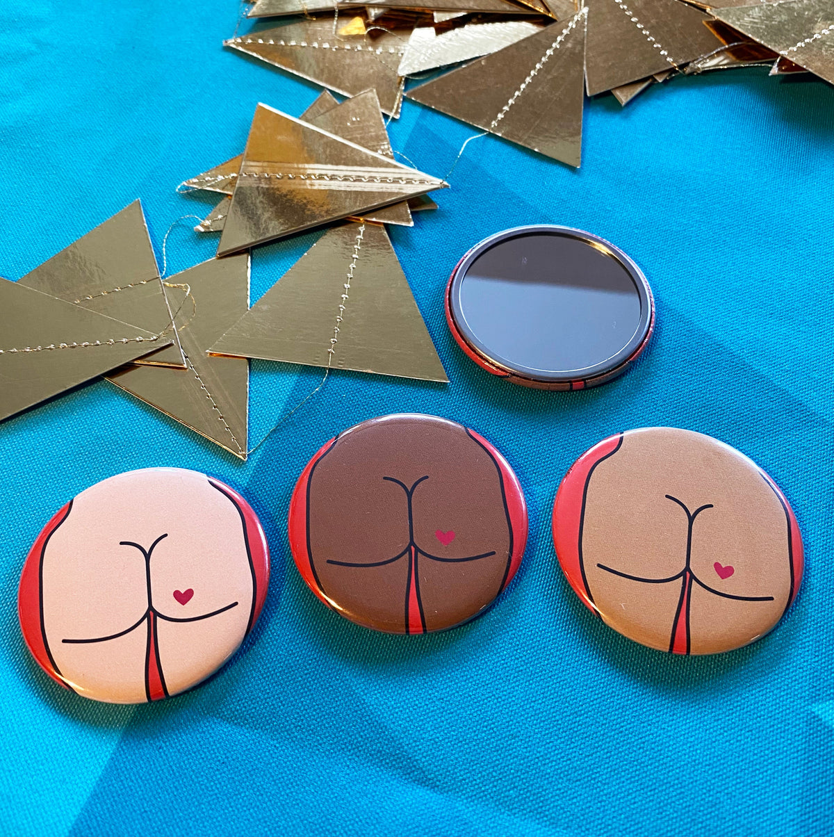 Four small mirrors on a blue background with gold triangles. One mirror is face up, the others are face down, showing their decorative backs that have cartoon illustrations of butts on them. 