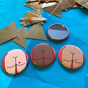 Four small mirrors on a blue background with gold triangles. One mirror is face up, the others are face down, showing their decorative backs that have cartoon illustrations of butts on them. 