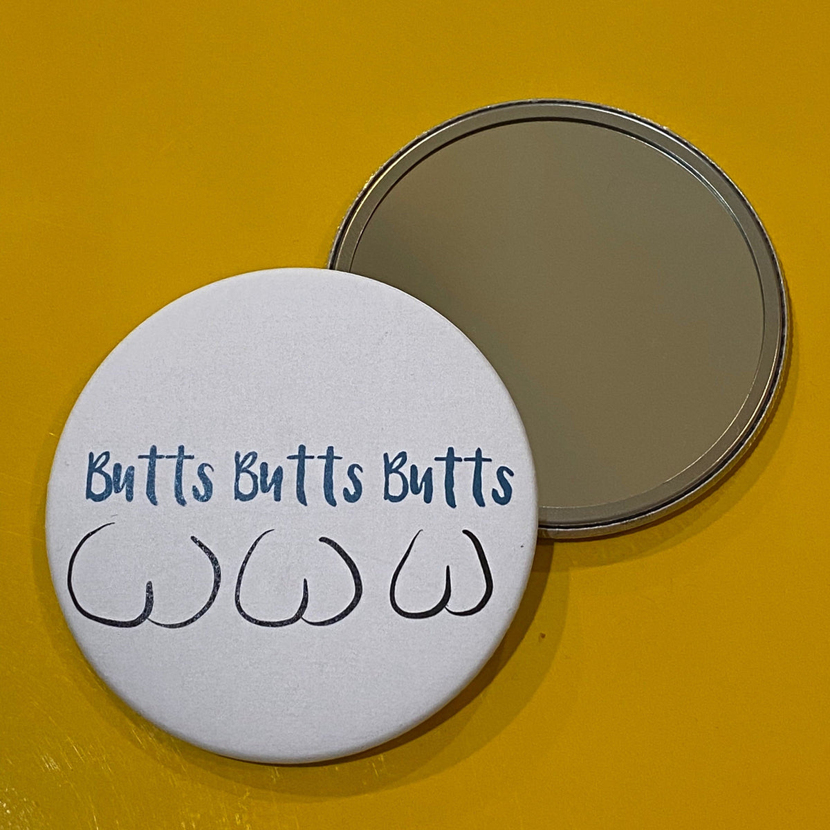 Two mirrors on a yellow background, one face up showing its reflective side, and one face down showing its decorative side, which has an illustration of three butts and text that says "Butts Butts Butts"