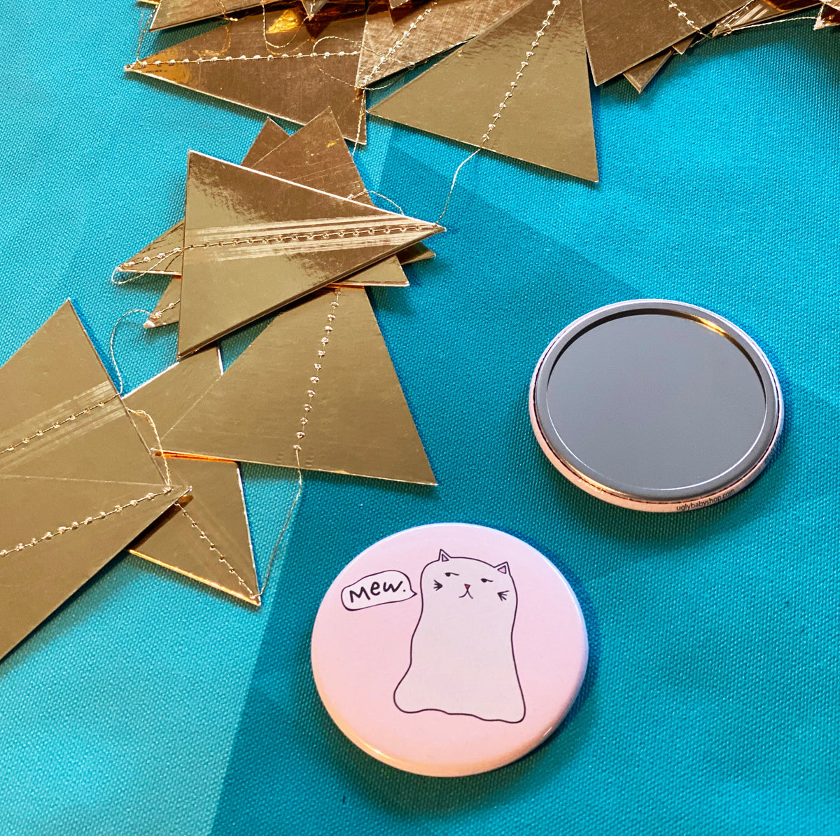 Two mirrors on a blue fabric surface with gold paper triangles. One is face up, showing its reflective surface, and the other is face down, showing a design of a ghost cat with a speech bubble that says "mew."