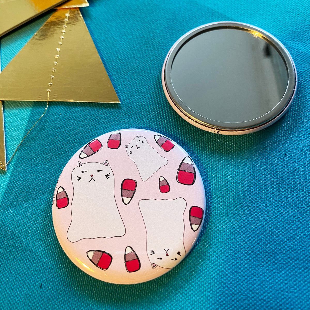 Two mirrors on a blue fabric background, one face up showing its reflective face, and the other face down showing a design of ghost cats surrounded by pink candy corn. 