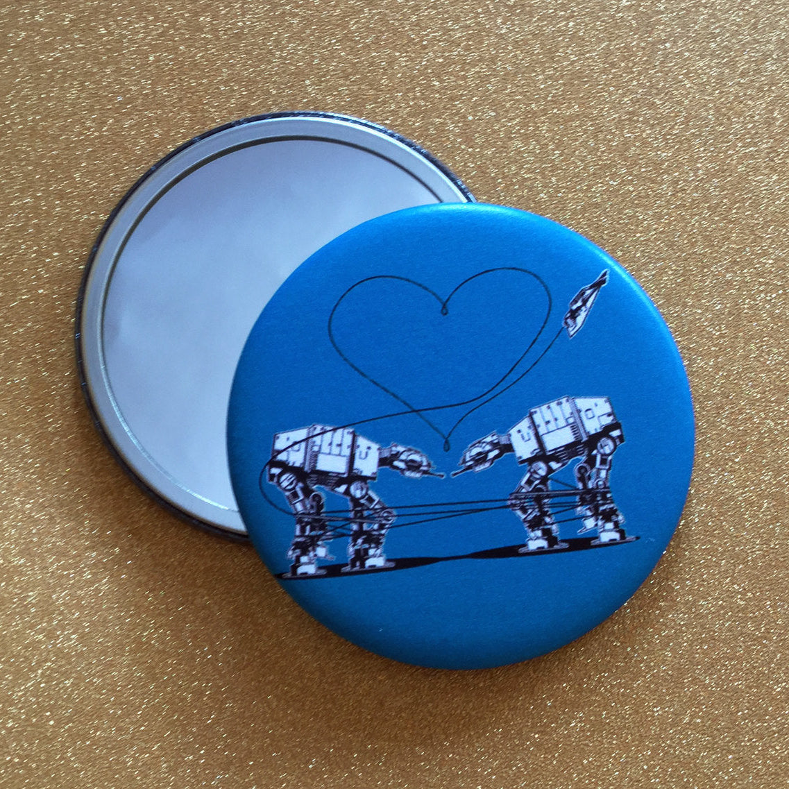 Two mirrors against a glittery gold surface. One is face up, showing its reflective surface, and the other is face down, showing a design of two AT-ATs from Star Wars against a blue background. 