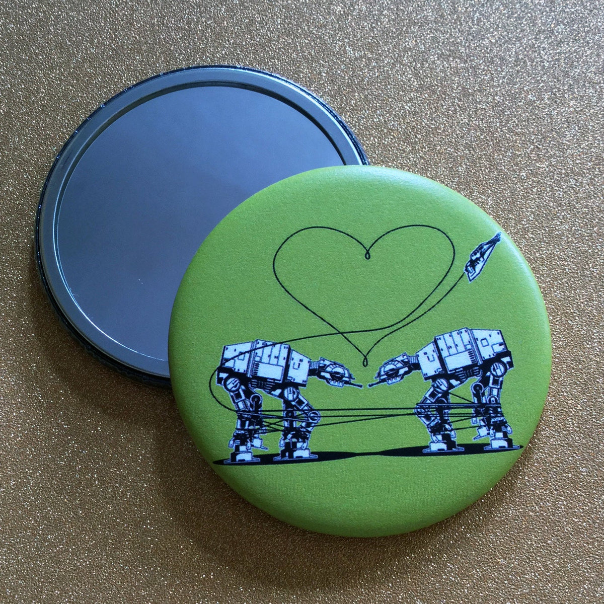 Two mirrors against a glittery gold background. One is face up, showing its reflective surface, and the other is face down, showing a design of two AT-ATs from Star Wars against a green background. 