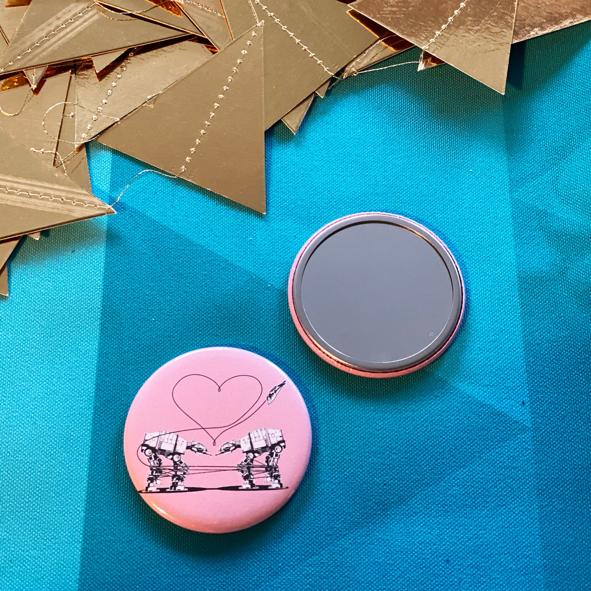 Two mirrors on a blue fabric background with gold paper triangles. One is face up, showing its reflective surface, the other is face down showing an illustration of two AT-ATs from Star Wars on a pink background. 