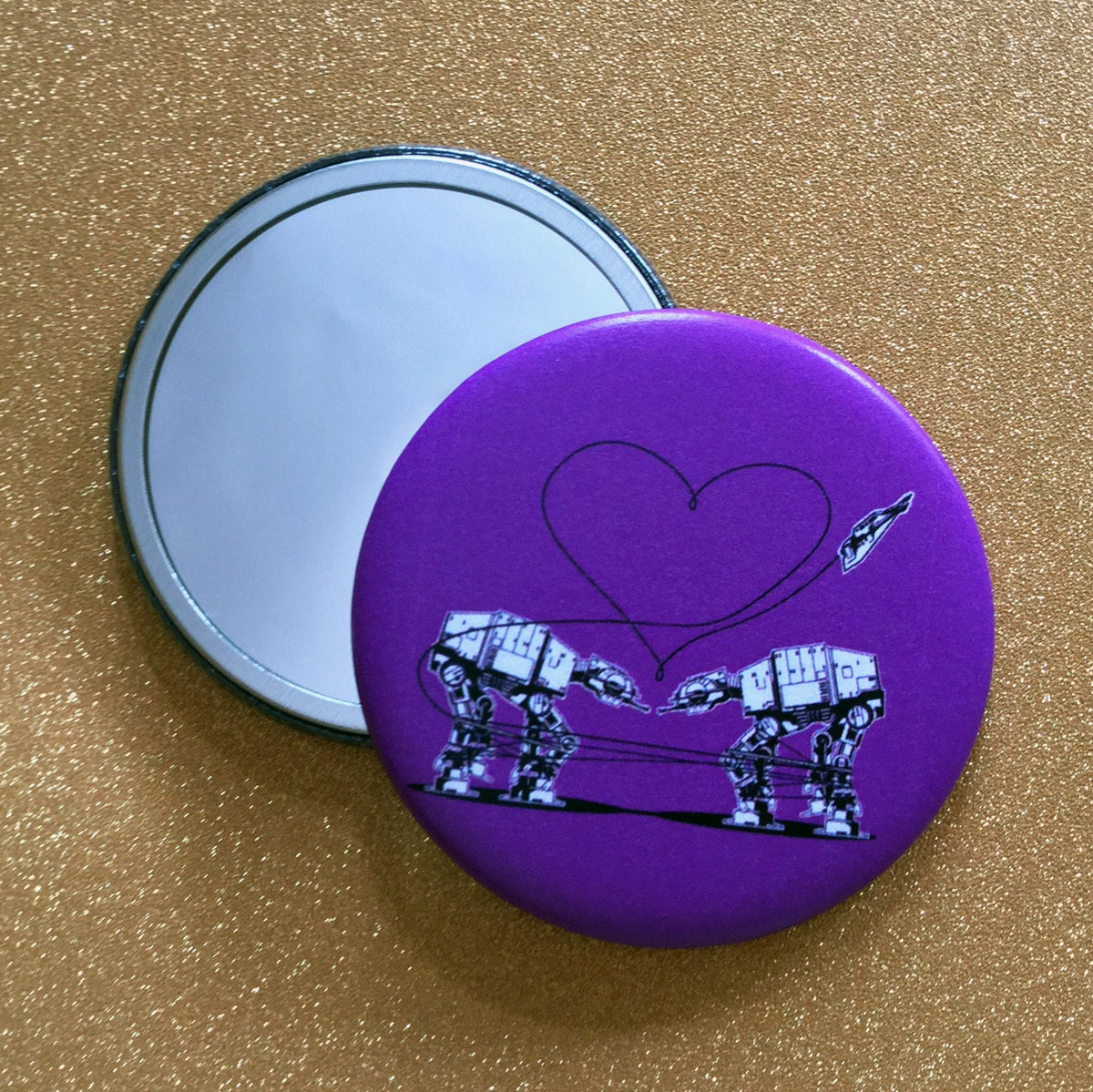 Two mirrors on a gold glittery background. One mirror is face-up, showing its reflective side. The other is face down, showing an illustration of two AT-ATs from Star Wars on a purple background. 