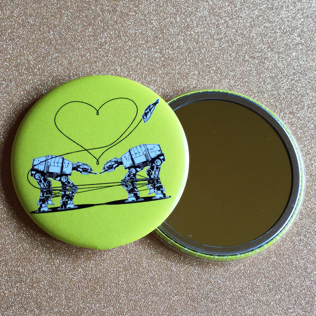 A round yellow mirror with an illustration of two AT-ATs from Star Wars with a heart above them, against a glittery gold background.