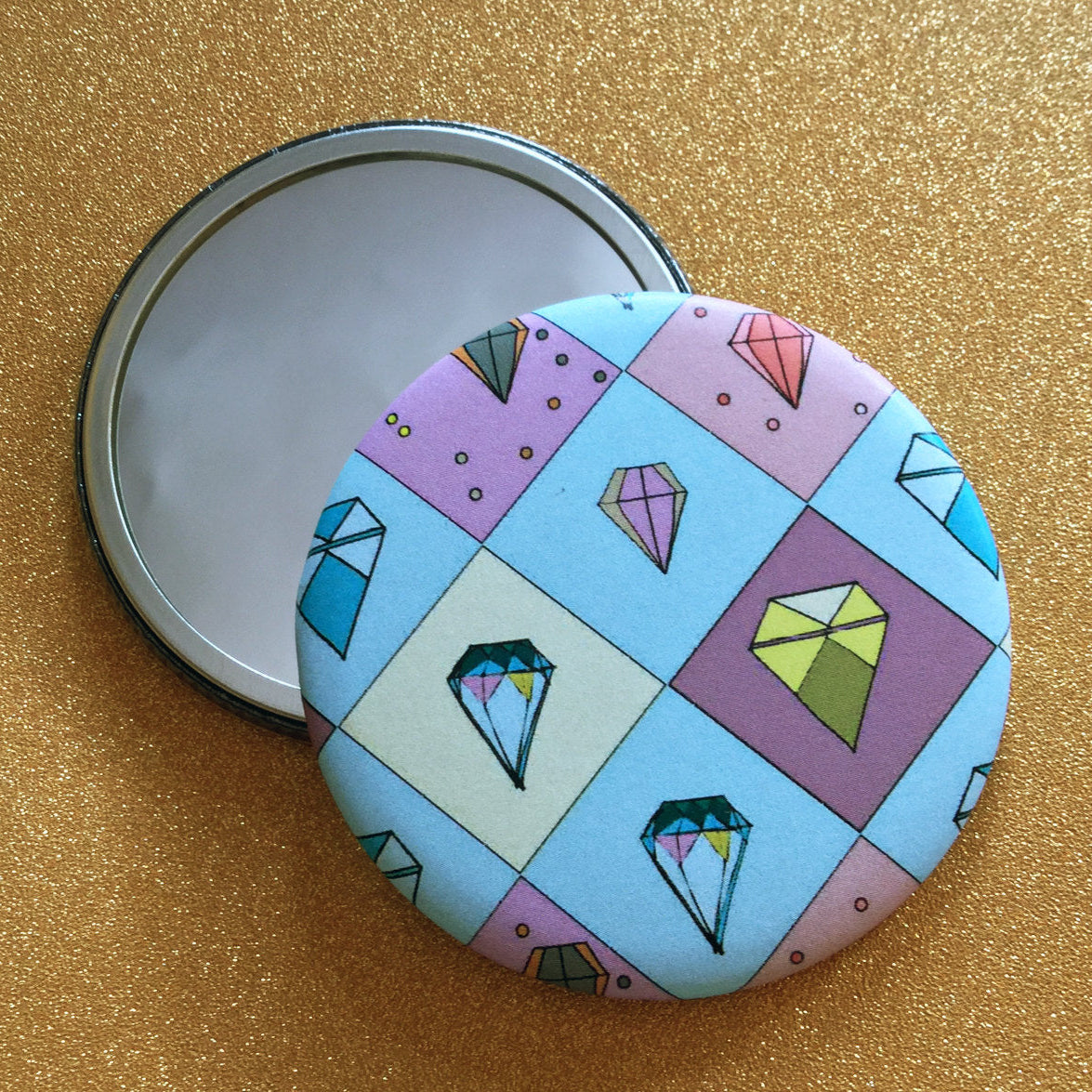 A small round mirror with a diamond-shaped pattern with different colored gemstones on it, against a glittery gold background. 