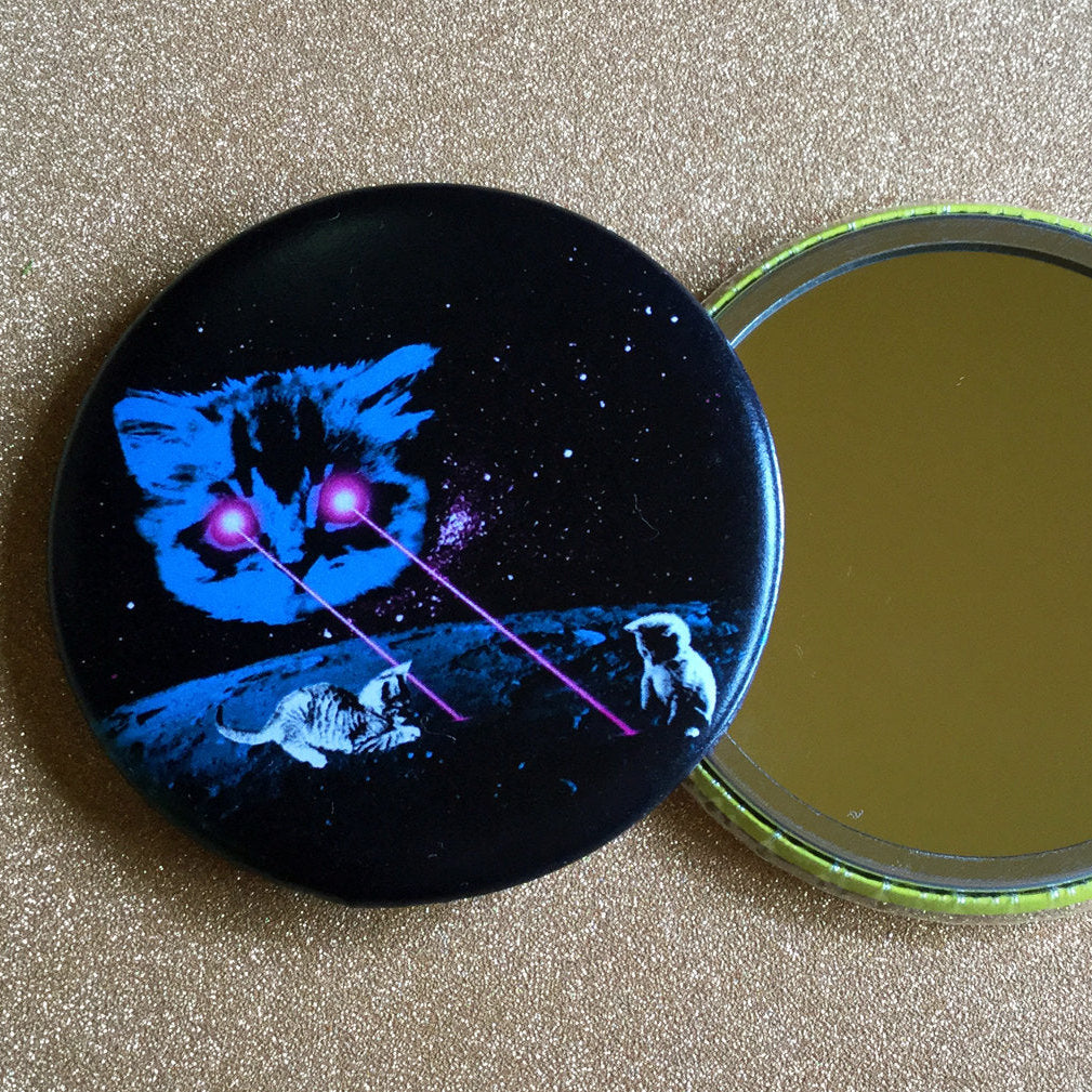 A small round mirror with a design of a cat shooting lasers out of its eyes at two smaller cats with a galaxy background. 