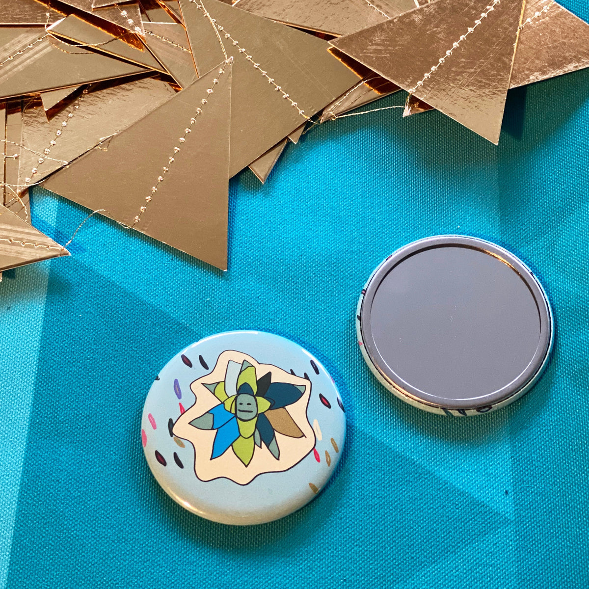 Round mirror with a succulent illustration against a blue fabric background with gold paper triangles.