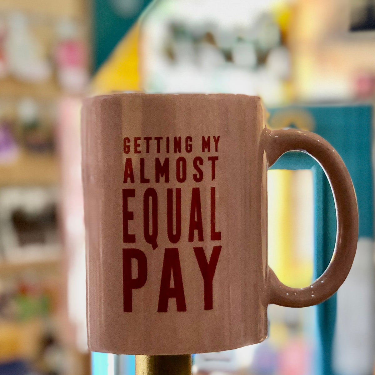 Pink coffee mug with red text that reads "GETTING MY ALMOST EQUAL PAY" against a blurred background. 