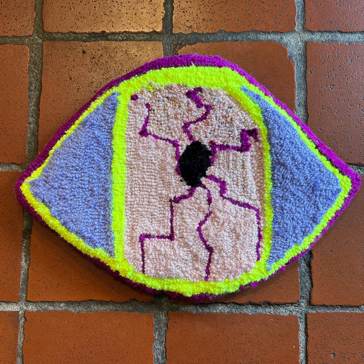 A piece of tufted artwork in the shape of an eyeball with pink, purple, lilac, and neon yellow colors against a brick background. 