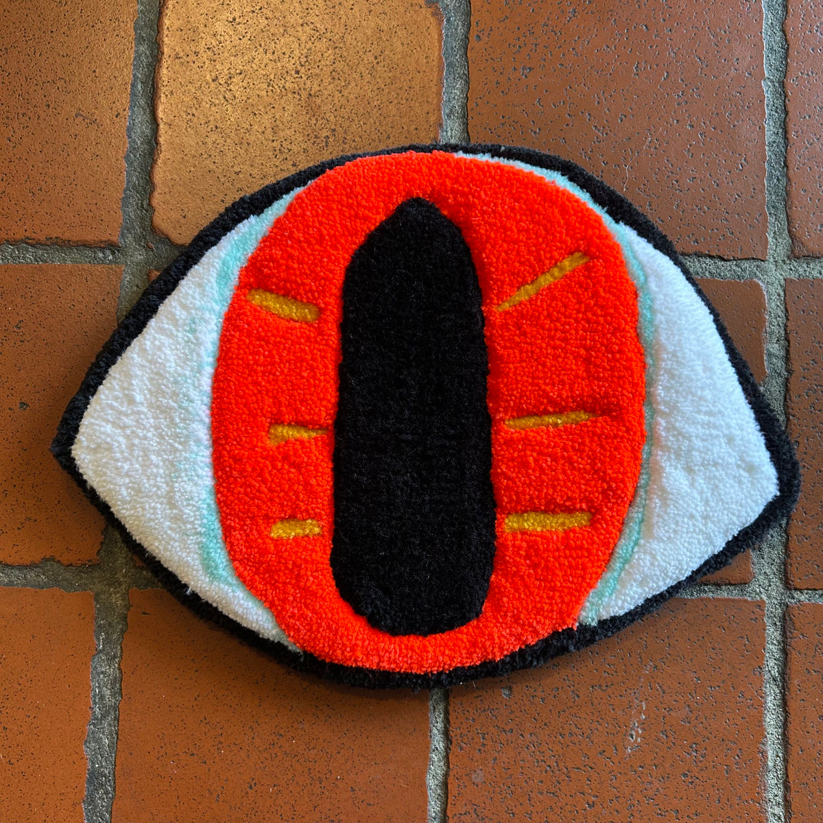 A tufted art piece in the shape of an eye with a black outline and pupil, and an orange iris with yellow stripes against a brick background. 