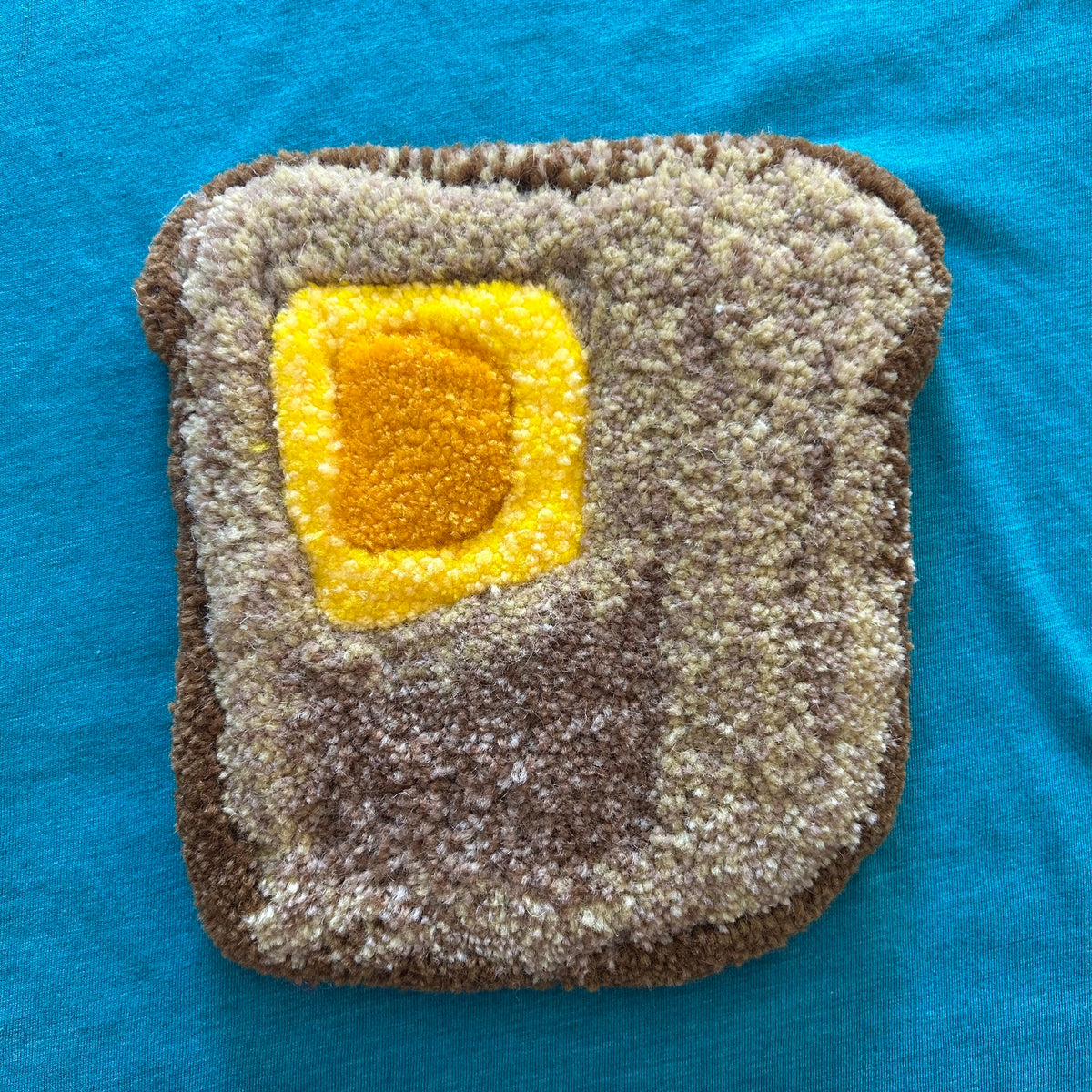 A piece of tufted artwork in the shape of a piece of toast with a yellow pat of butter on top in front of a blue fabric background. 