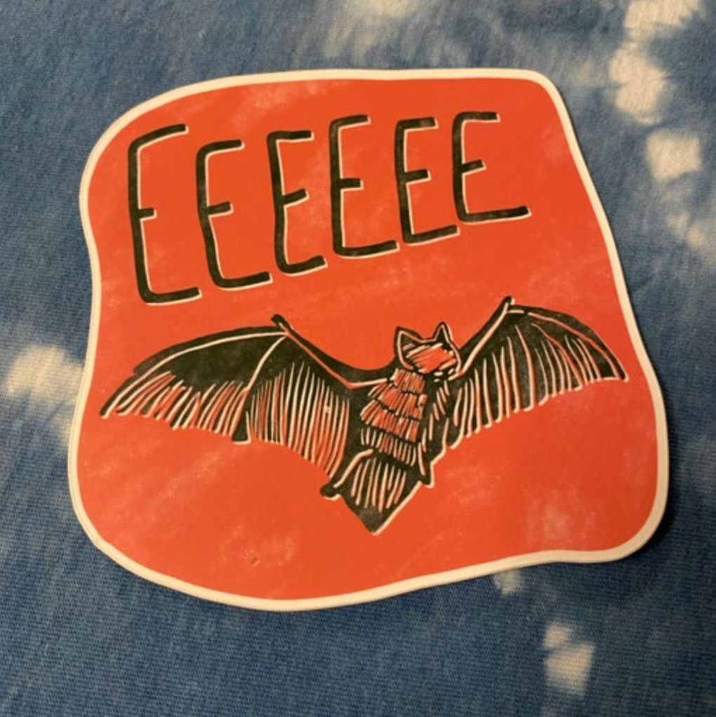 Sticker with a bat illustration and 'EEEEE' text on an orange background, placed on a denim surface.