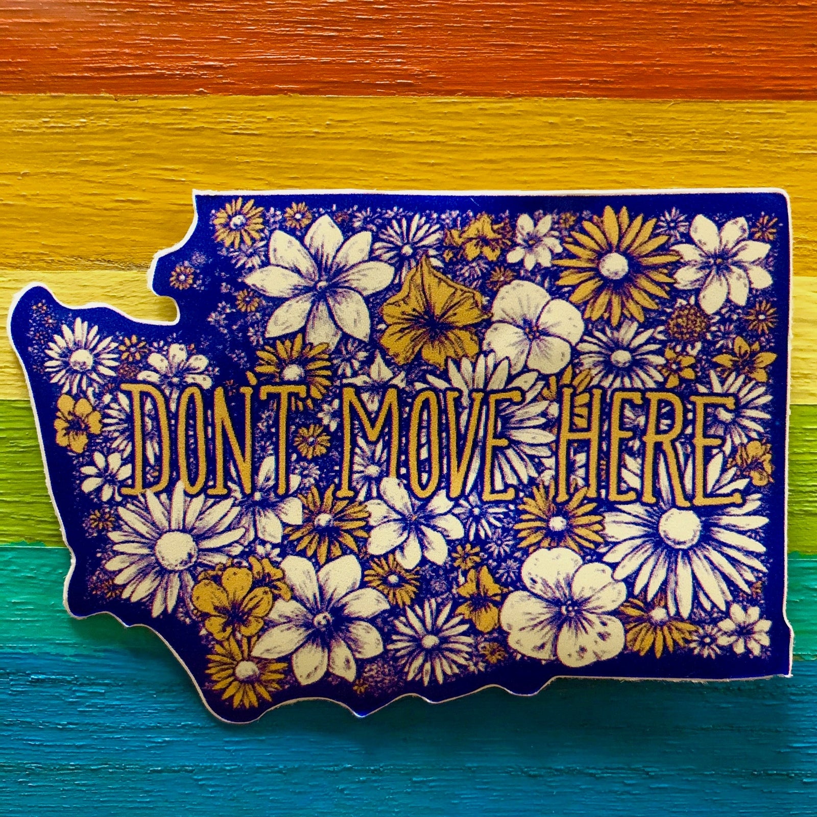 Washington state-shapes sticker with flowers and text that says "DON'T MOVE HERE" against a painted rainbow background
