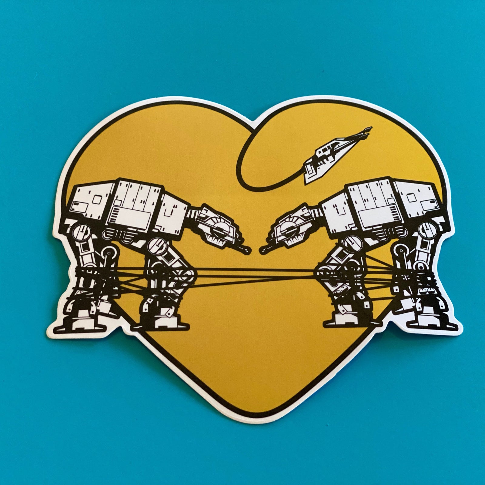 Yellow heart shaped sticker with two robots on a blue background. 