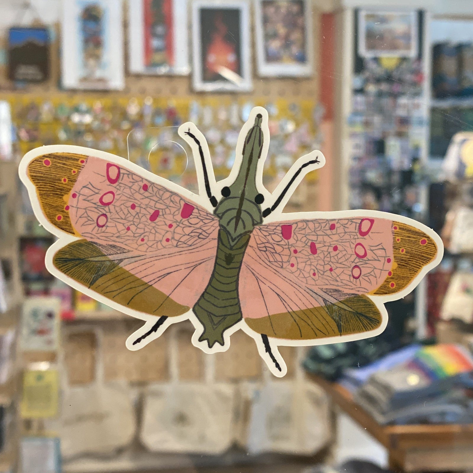 A sticker of a moth with pink wings against a blurred background. 