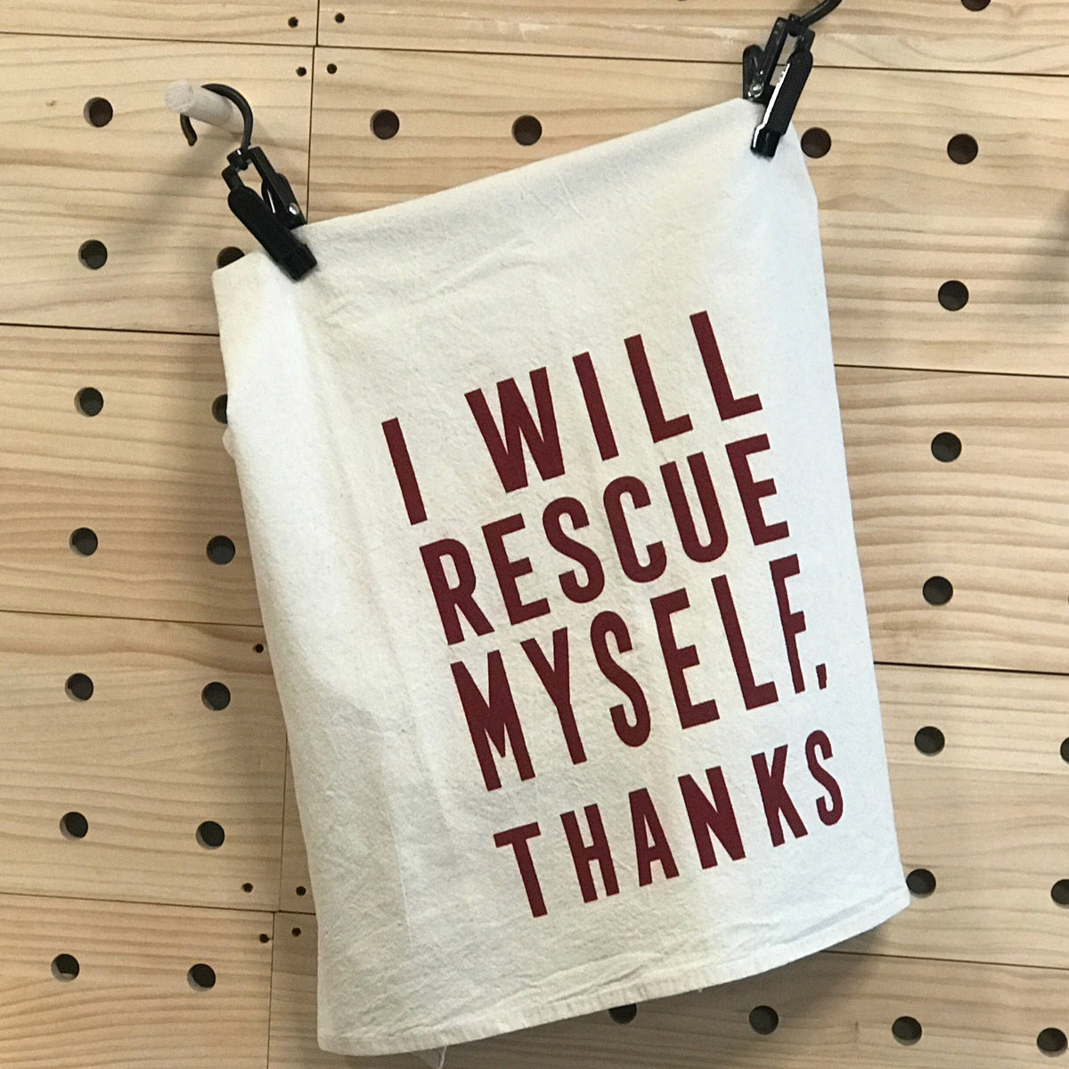 A natural color flour sack towel is folded so you can read the words, "I Will rescue myself, thanks" in screenprinted maroon lettering. It is hanging against a wood pegboard wall. 