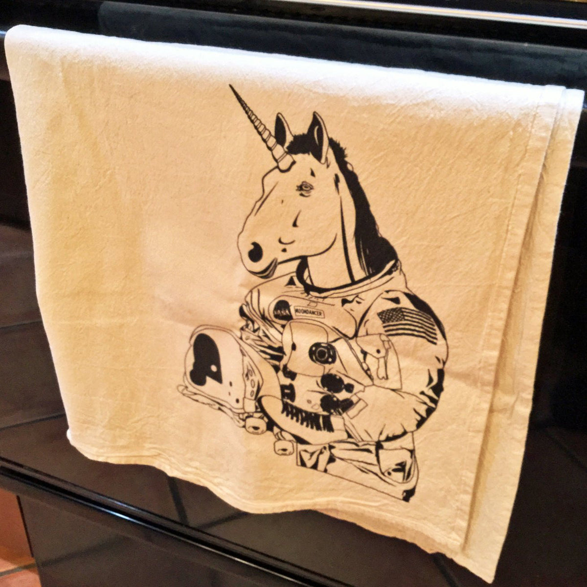 A natural color kitchen towel screenprinted with black ink. It has an image of a unicorn wearing rollerskates and holding an astronaut helmet. His horn will obviously not fit inside the helmet. The towel is hanging on a stove handle.