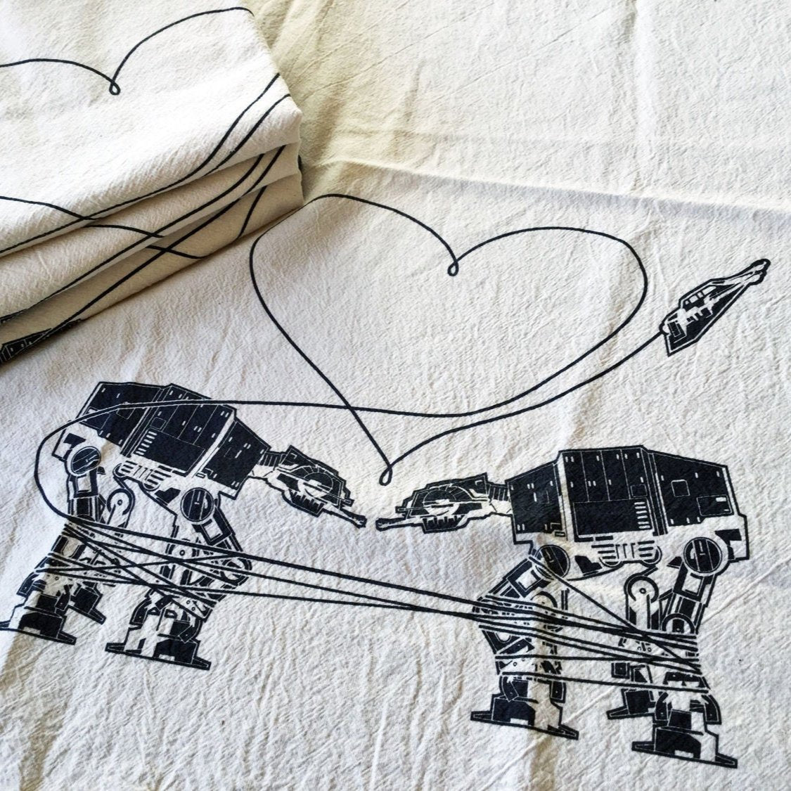 Two All Terrain Armoured Transports are gazing lovingly into each other's eyes while a heart forms above them. The image is printed on a natural color kitchen towel with black ink.