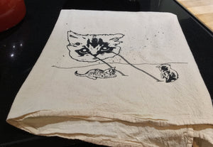 This natural color kitchen towel has a space cat shooting lasers at tiny kitties. The kittens are playing in the lasers. The towel is printed with black ink and is folded on top of a black stove. 