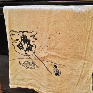 This natural color kitchen towel has a space cat shooting lasers at tiny kitties. The kittens are playing in the lasers. The towel is printed with black ink and is hanging on a black oven door.